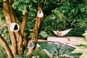 Drie lichtgrijze, organisch gevormde vogelhuisjes van Elho bevestigd aan een boom bij een zwembad in een tuin.