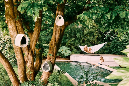 Drie lichtgrijze, organisch gevormde vogelhuisjes van Elho bevestigd aan een boom bij een zwembad in een tuin.