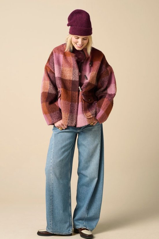 Model draagt een donkerroze en bruin geruit wollen jack, donkerroze muts, wijde jeans en platte schoenen.