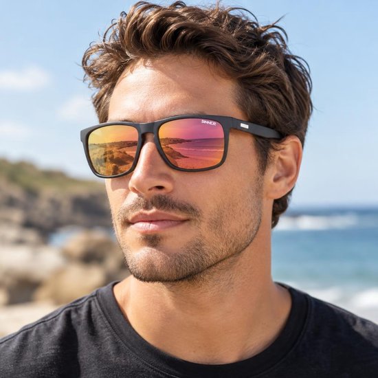 Man draagt zwarte SINNER zonnebril met reflecterende oranje-roze lenzen bij de zee.