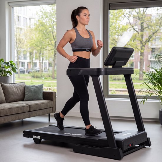 Vrouw met een VirtuFit hartslagmeter om haar borst, gekleed in sportkleding, traint op een loopband in een woonkamer.