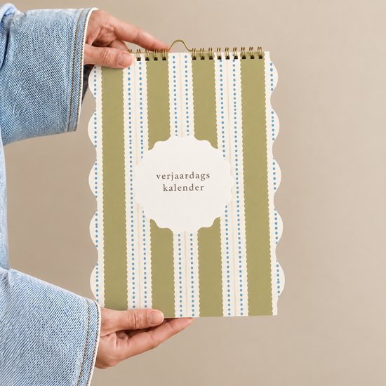 Verjaardagskalender met verticale groen-beige strepen en blauwe stippen. Rand golvend. Vastgehouden door handen in spijkerstof.