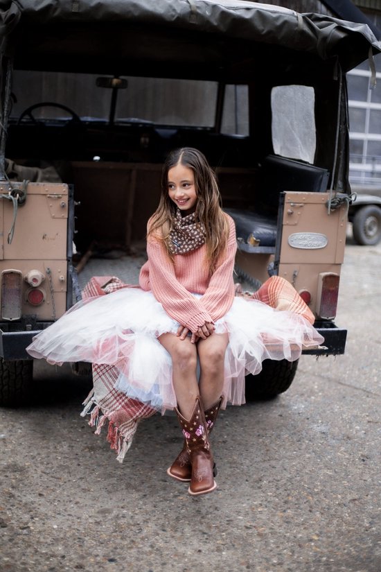 Meisje zittend op de achterkant van een auto, draagt een roze trui, witte tule rok en bruine cowboylaarzen met bloemen.