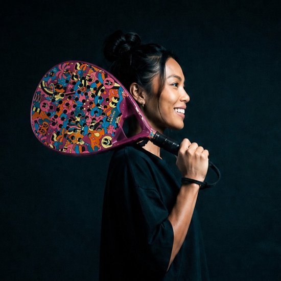 Vrouw met een padelracket met een kleurrijk, cartoonachtig patroon in felroze, oranje, blauw en geel. Padelracket wordt op haar schouder gedragen.