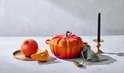Oranje Le Creuset pompoenvormige gietijzeren braadpan met zilverkleurige steel, naast een verse pompoen.