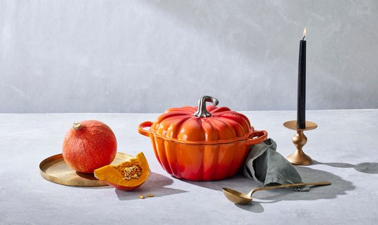 Oranje Le Creuset pompoenvormige gietijzeren braadpan met zilverkleurige steel, naast een verse pompoen.