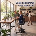 Een vrouw in een donkerblauwe blazer en witte broek zit op een zwarte bureaukruk met voetsteunring. Ze praat met een man aan een houten tafel. Er staat "Zoals een barkruk maar dan actief" op de muur.
