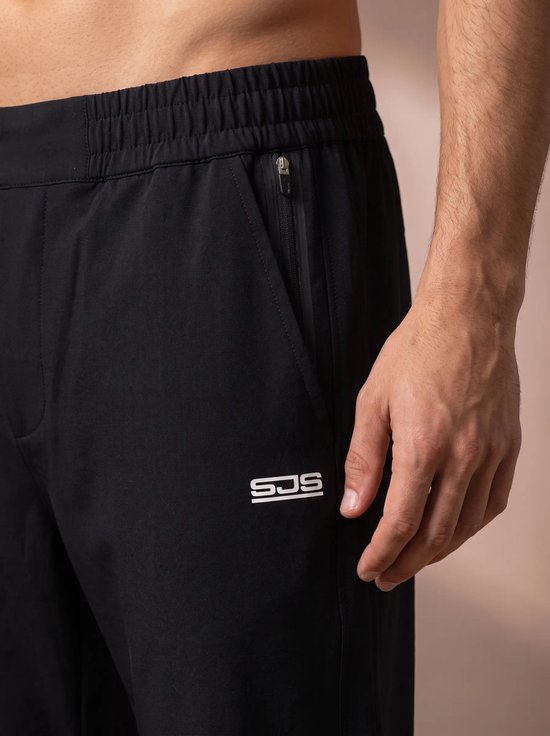 Close-up van een zwarte sportbroek met ritssluiting en het 'SJS' logo, gedragen door een man.