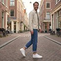 Man met een beige jack, blauwe jeans en witte sneakers met witte sokken, lachend in een straat.