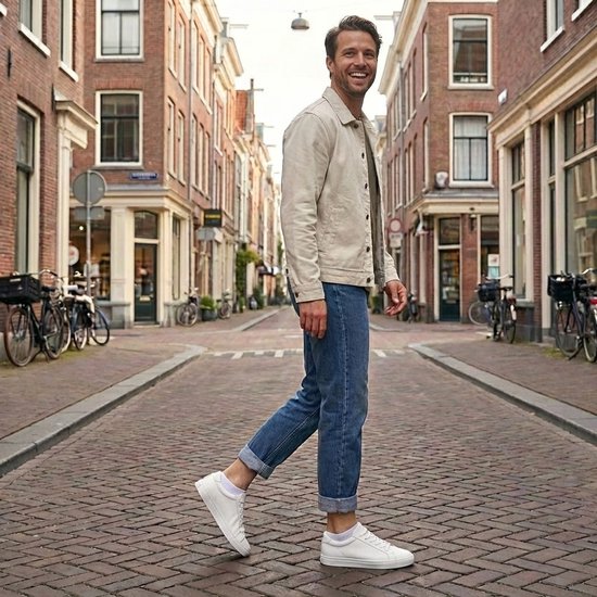 Man met een beige jack, blauwe jeans en witte sneakers met witte sokken, lachend in een straat.