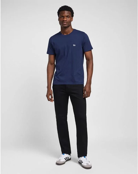 Volledig zicht van een man in een marineblauw T-shirt en zwarte jeans, met witte sneakers met strepen.