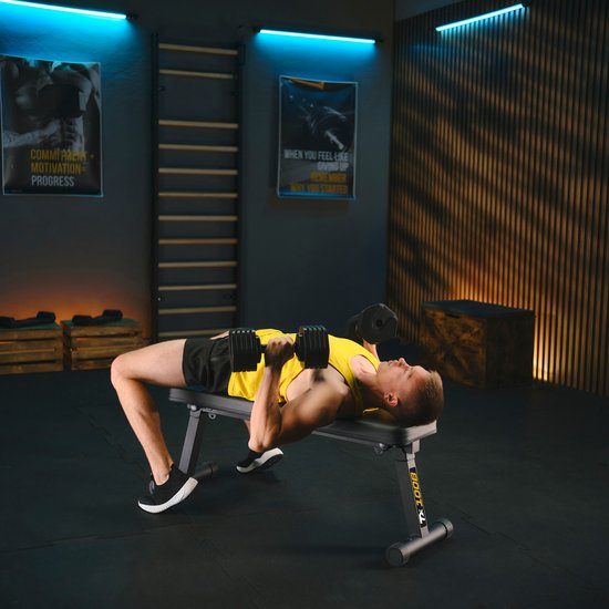 Een man in een gele tanktop en zwarte shorts doet een dumbbell-oefening op een zwarte trainingsbank.
