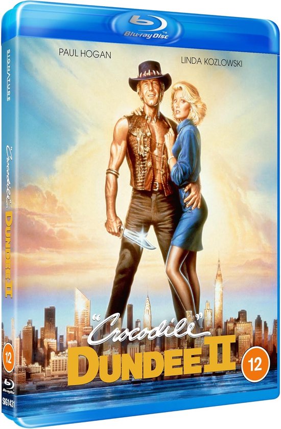 Blu-ray hoes van Crocodile Dundee II. Paul Hogan in cowboykleding met mes, Linda Kozlowski in blauwe jurk, met stadsgezicht.