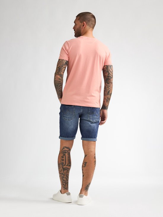 Man, staand, in een roze T-shirt, spijkerbroek met opgerolde zoom en witte sneakers.