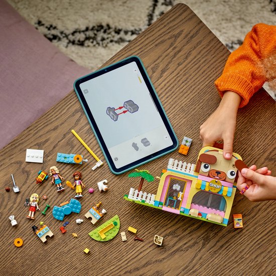 Mains d'enfant construisant avec des briques LEGO Friends sur une table en bois. Une tablette affiche une instruction de montage. Une animalerie est prête.