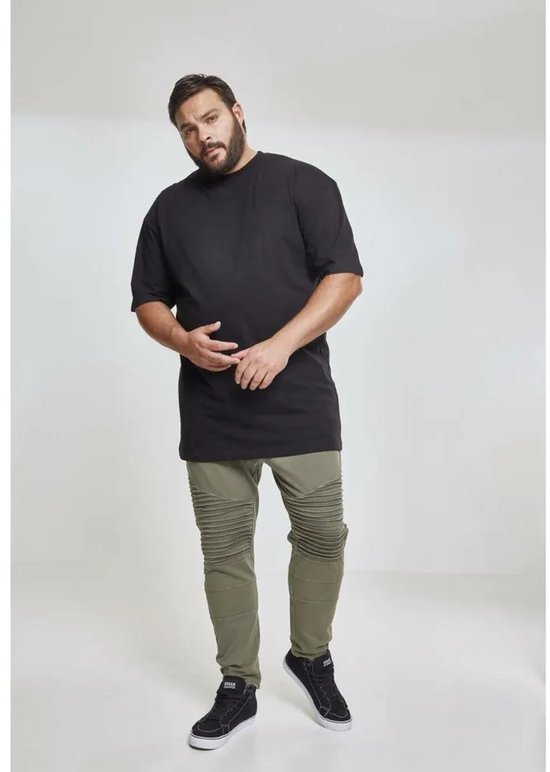 Een volslanke man staat frontaal in een zwart oversized T-shirt en een groene broek met geribbelde knieën en zwarte sneakers.