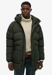 Een man draagt een lange, donkergroene, gewatteerde parka met capuchon, ritssluiting en knopen.