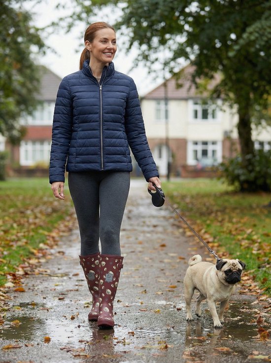 Een vrouw wandelt met een hond op een herfstig, nat pad. Ze draagt een donkerblauwe jas, grijze legging en bordeauxrode regenlaarzen met mopshondenprint.