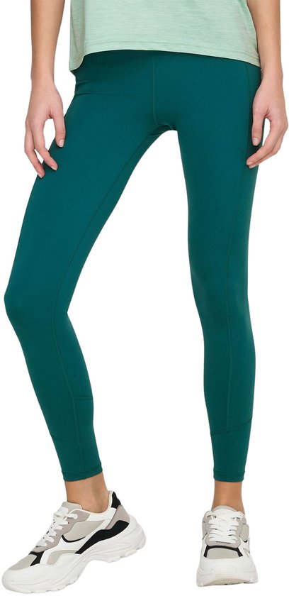 Model in een donkergroene legging met zakken aan de zijkant, sneakers en lichtgroen shirt.