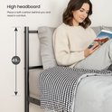 Une femme lit un livre dans un lit avec une haute tête de lit en métal noir, couverte d'une couette grise et d'une couverture à carreaux.