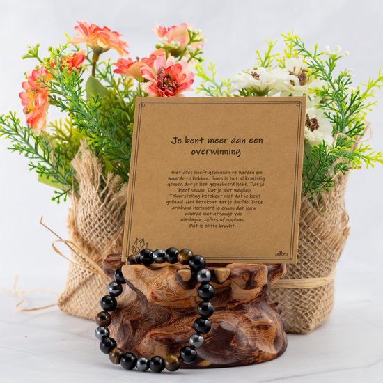 Armband met onyx, tijgeroog en hematiet kralen op een houten display, voor een kaart met tekst en bloemen.