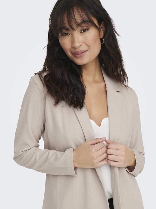 Vrouw met lange, golvende donkere haren in een lange, beige blazer, met de handen op de voorkant.