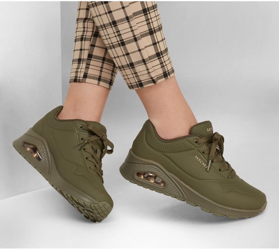 Model draagt legergroene Skechers Uno damessneakers met geruite broek. Zichtbaar luchtkussen in de zool.