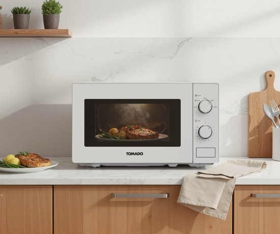 Witte Tomado solo magnetron op aanrecht, met opengewerkte deur waarin een bord met eten stoomt. Links een bord met vlees, rechts keukenaccessoires.