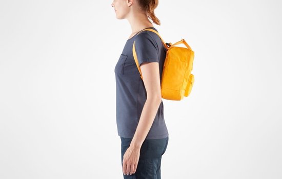 Een vrouw draagt de gele Kånken Mini rugzak aan haar rechter schouder, profiel.