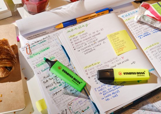 Groene en gele STABILO BOSS markers op een bureau vol aantekeningen, naast een croissant en een glas.