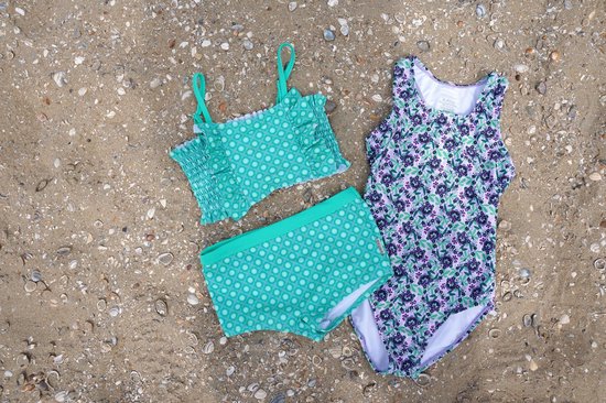 Paars en groen gebloemd badpak, mintgroene bikini met ruches en groene bikinibroekje met cirkelpatroon op zand.