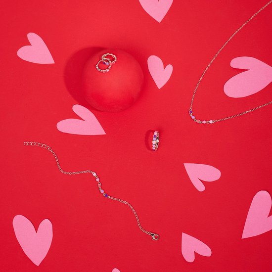 Zilverkleurige ketting, armband en twee ringen met paarse en roze hartjes, tegen een rode achtergrond met hartjes.