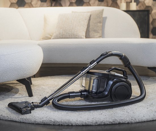 Aspirateur Tomado noir avec tuyau et embout sur un tapis doux, près d'un canapé blanc.
