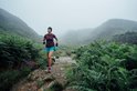 Vrouw rent op een bergpad in de mist, draagt een paarse shirt, blauwe short en roze sokken.