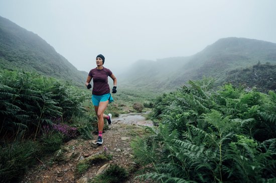 Vrouw rent op een bergpad in de mist, draagt een paarse shirt, blauwe short en roze sokken.
