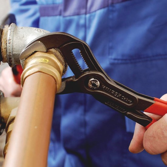 Een hand met Knipex Cobra tang met rode handvatten monteert een koppeling op een koperen buis.
