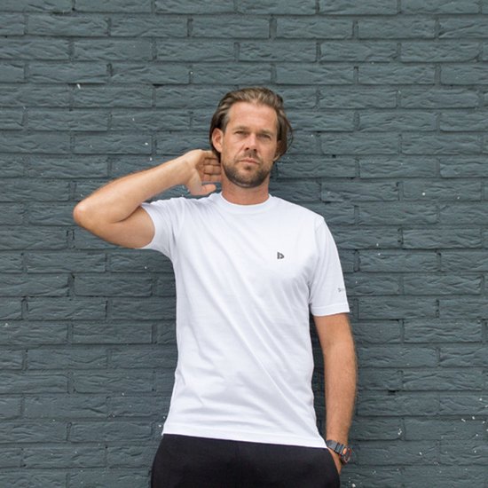 Man met witte Donnay t-shirt en zwarte broek, leunt tegen donkergrijze bakstenen muur.