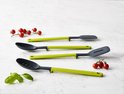 Ensemble de quatre ustensiles de cuisine : deux cuillères perforées, une spatule et une cuillère, avec des manches verts et gris foncé, avec des tomates et des herbes.
