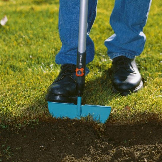 Man met blauwe jeans en zwarte schoenen gebruikt de turquoise graskantafsteker in het gras.