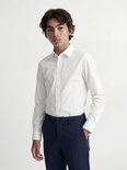 Un garçon porte une chemise blanche à manches longues et un pantalon bleu.