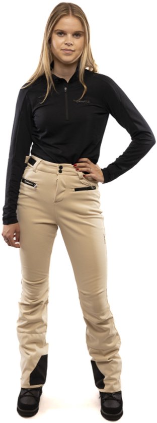 Model in een beige skibroek met zwarte top, staand met een hand op de heup.