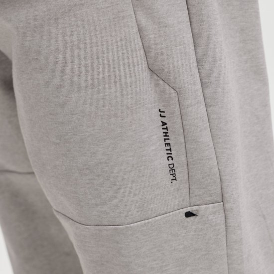Detail van grijze joggingbroek met tekst 'JJ ATHLETIC DEPT.' en een klein zwart logo.
