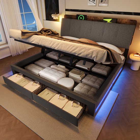 Grijze gestoffeerde bed met openklapbaar matras die toegang geeft tot opbergruimte met kleding in de open lades eronder. Het hoofdbord heeft LED-verlichting.