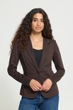 Vrouw draagt een bruine blazer en spijkerbroek, met handen gevouwen. Ze heeft krullend haar.