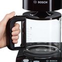 Hand die een zwarte Bosch koffiemachine pakt bij de zwarte handgreep van de glazen kan.