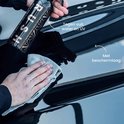 Hand met zwarte handschoen poetst glanzende zwarte auto met beige doek. Fles RUSH Spray Wax naast de auto. Tekst: Tegen vuil, water en UV. Met beschermlaag.