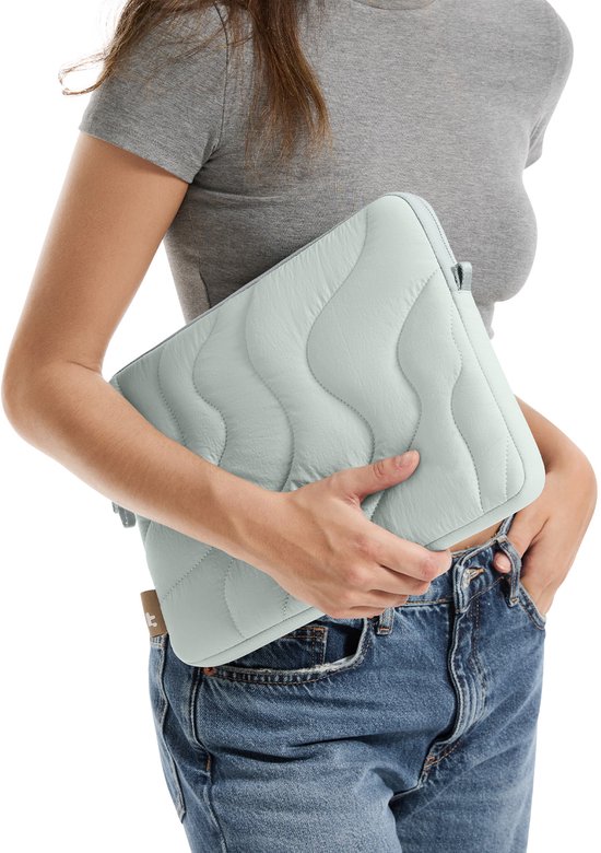 Une femme en t-shirt gris et jean tient une housse d'ordinateur portable bleu clair, matelassée, avec des surpiqûres ondulées.