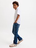 Jongen loopt in witte t-shirt en blauwe jeans met bruine sneakers, hand in zak.