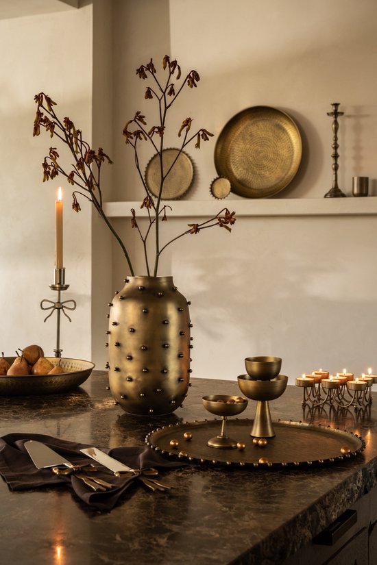 Gouden vaas met spikes en gedroogde takken, staat op een donkere tafel met gedekte tafel met messing dienblad, kandelaar en kaarsen.
