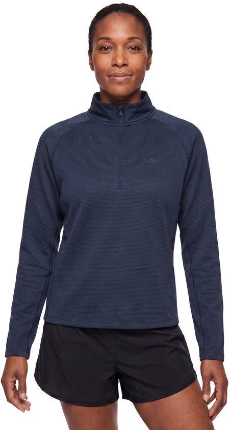Vrouw staat in de studio, draagt een marineblauwe, lange mouwen top met halve rits en zwarte sportshort.